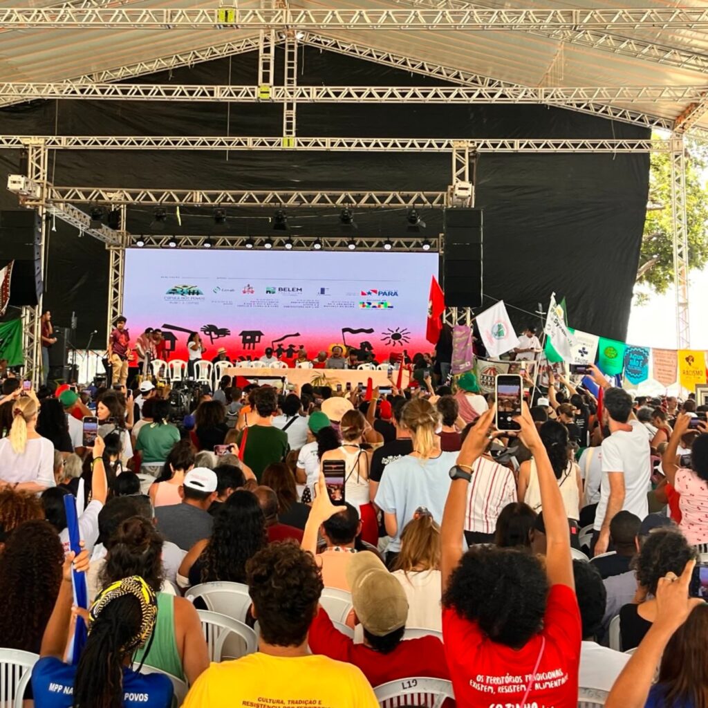 Público acompanha ato público em frente a palco ao ar livre, com bandeiras erguidas e pessoas registrando o momento com celulares