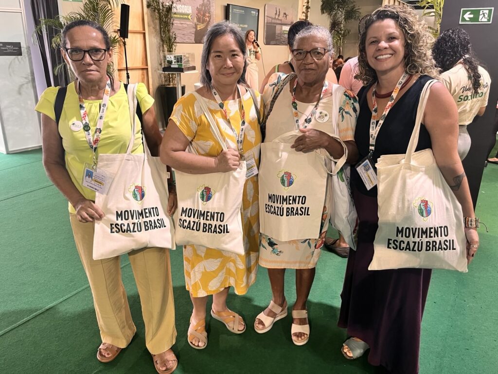 Quatro mulheres posam juntas em evento, usando crachás e segurando bolsas do Movimento Escazú Brasil.