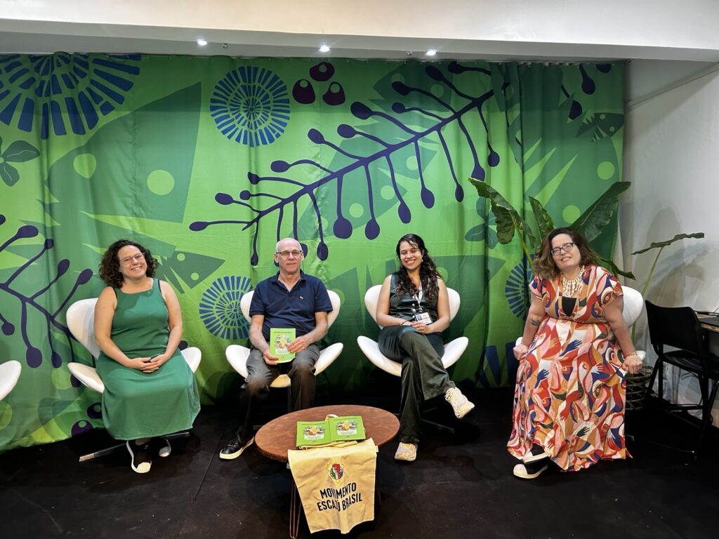 Quatro pessoas sentadas em cadeiras participam de conversa em palco, diante de painel verde com grafismos de folhas e com uma mesa de centro contendo folders e a bolsa do Movimento Escazú Brasil.