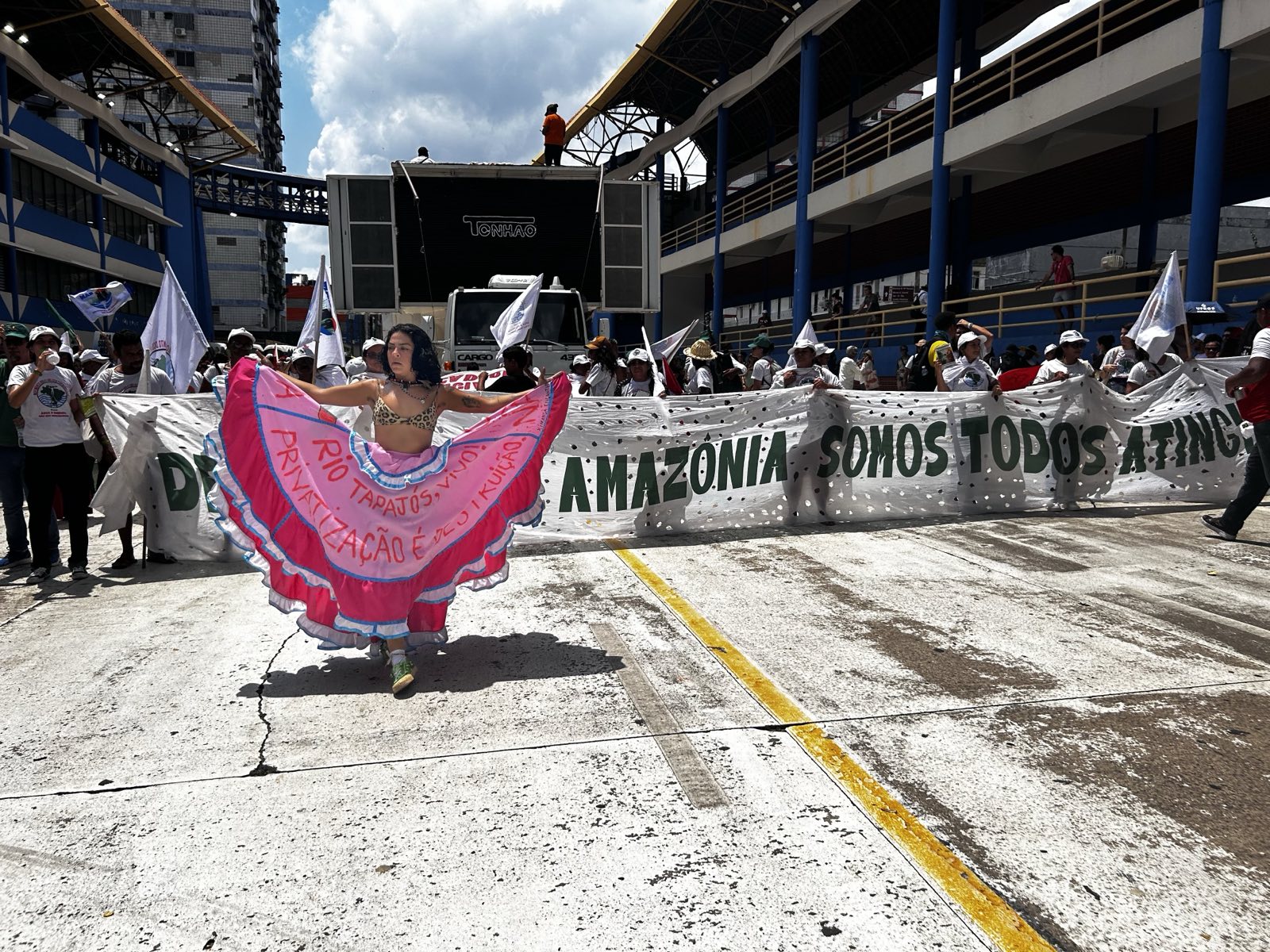 Ato em espaço urbano: mulher dança com saia colorida à frente de manifestantes que seguram faixa com a frase “Amazônia, somos todos atingidos”. Ato em espaço urbano: mulher dança com saia colorida à frente de manifestantes que seguram faixa com a frase “Amazônia, somos todos atingidos”.