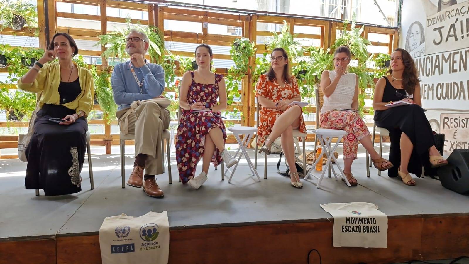Seis pessoas sentadas em um palco participam de mesa de debate, com plantas ao fundo e bolsas do Movimento Escazú Brasil na frente do palco. Seis pessoas sentadas em um palco participam de mesa de debate, com plantas ao fundo e bolsas do Movimento Escazú Brasil na frente do palco.