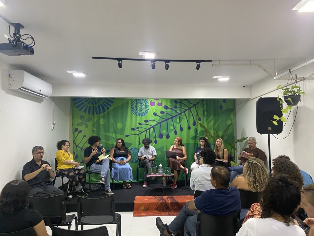 Público assiste a mesa de debate com várias pessoas sentadas em palco, diante de painel verde com grafismos naturais, em espaço fechado.
