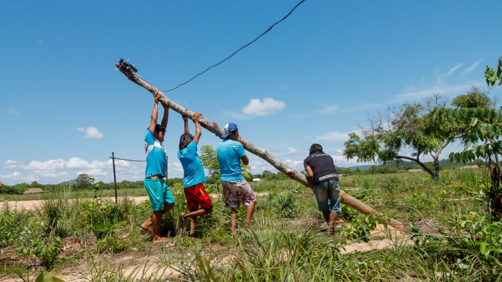 Grupo de homens em área rural trabalhando juntos para erguer um poste de madeira com roteador de internet para levar conectividade à comunidade.